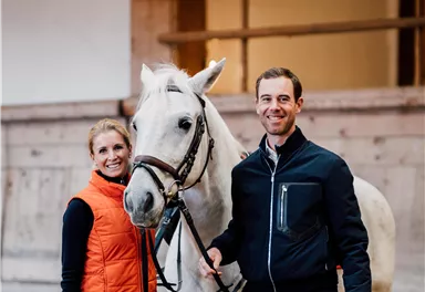 Eine Frau und ein Mann stehen mit einem weißen Pferd in einem Reitstall. Der Mann trägt eine dunkle Jacke und die Frau eine orange Weste.