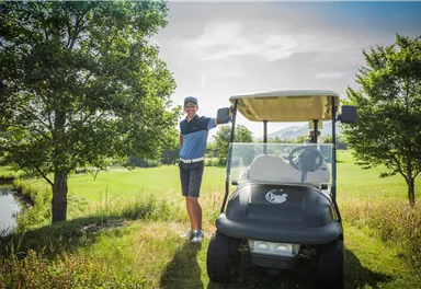Ein Mann steht neben einem Golfwagen auf einem Golfplatz. Im Hintergrund sind Bäume und eine grüne Wiese zu sehen.