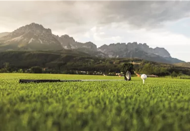 Ein Golfplatz mit grünem Gras und einem Golfball auf einem Tee. Im Hintergrund sind beeindruckende Berge und ein bewölkter Himmel zu sehen.