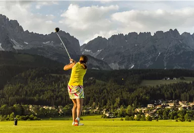A person is playing golf on a beautiful course, surrounded by mountains. The sky is partly cloudy and the landscape is green and well-maintained.
