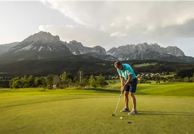 A golfer putts on a green course with picturesque mountains in the background. The landscape is calm and idyllic.