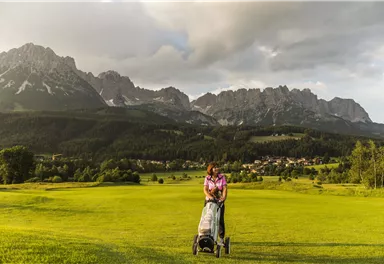A woman with a golf cart is standing on a green golf course. Impressive mountains and a cloudy sky can be seen in the background.