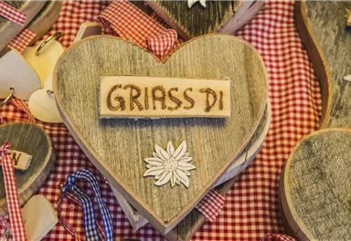 Ein hölzernes Herz mit der Aufschrift "GRIASS DI" und einer dekorativen Blume. Der Hintergrund ist mit kariertem Stoff dekoriert.