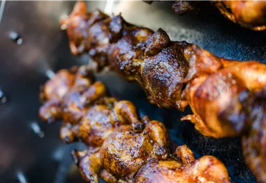 Ein köstlicher Spieß mit gegrilltem Hähnchen, das goldbraun und knusprig aussieht. Der Hintergrund zeigt eine Grillfläche mit weiteren Hähnchenstücken.