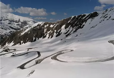 Eine malerische Berglandschaft mit schneebedeckten Hügeln und kurvenreichen Straßen. Der Himmel ist klar und blau, was eine atemberaubende Aussicht bietet.