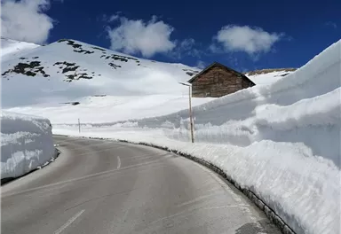Eine schneebedeckte Straße umgeben von hohen Schneewänden. Im Hintergrund steht ein kleines Holzhaus unter einem blauen Himmel mit einigen Wolken.