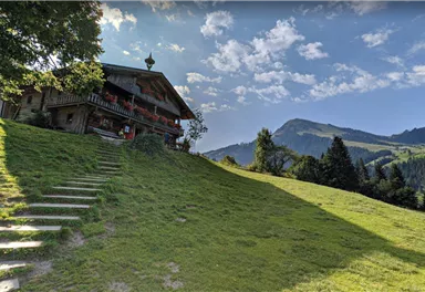 Ein malerisches Landhaus auf einer grünen Wiese mit sanften Hügeln im Hintergrund. Der Himmel ist klar und blau, mit einigen Wolken.
