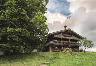 Ein traditionelles Holzhaus steht auf einem sanften Hügel, umgeben von Bäumen. Der Himmel ist bewölkt, was eine ruhige Atmosphäre schafft.