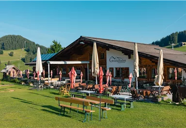 A cozy restaurant with an outdoor terrace, surrounded by meadows and mountains. Sunshades provide shade for the guests.