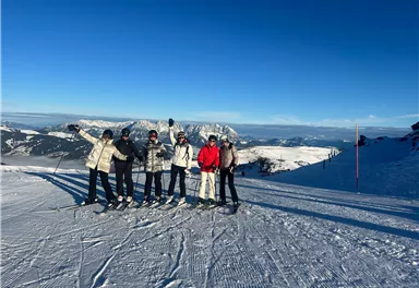 Fünf Skifahrer stehen auf einer verschneiten Piste. Im Hintergrund sind Berge und ein klarer blauer Himmel zu sehen.
