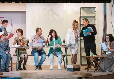 Eine Theateraufführung mit mehreren Schauspielern auf der Bühne. Die Darsteller sind in lebhaften Kostümen und interagieren an einem Tisch.
