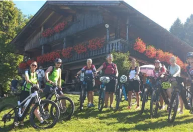 Eine Gruppe von Radfahrern steht lächelnd auf einer Wiese vor einem traditionellen Holzhaus. Im Hintergrund sind Bäume und blühende Pflanzen zu sehen.