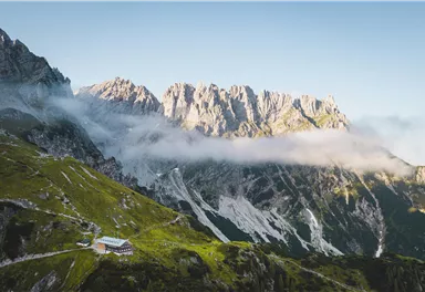 Eine beeindruckende Berglandschaft mit hohen Gipfeln und wolkenbedeckten Hängen. Im Vordergrund ist eine gemütliche Hütte zu sehen.