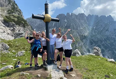Eine Gruppe von Menschen steht vor einem Bergkreuz in den Bergen. Sie lächeln und posieren fröhlich für das Foto.