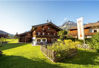 Ein traditionelles Bauernhaus in den Bergen mit schneebedeckten Gipfeln im Hintergrund. Die Umgebung ist grün und einladend, ideal für ländliche Erholung.