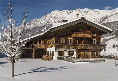 Ein malerisches Chalet im Schnee mit Holzverkleidung. Im Hintergrund sind majestätische Berge und ein klarer blauer Himmel zu sehen.