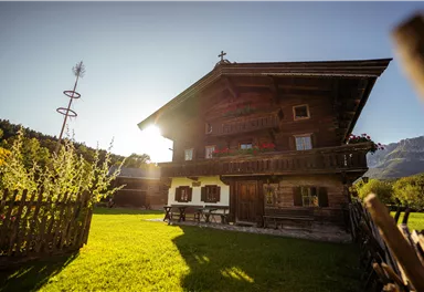 Eine gemütliche Stube mit Holzwänden und zwei Fenstern. Ein runder Tisch mit einer weißen Tischdecke steht in der Mitte, umgeben von Bankauflagen.