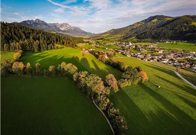Zwei Wanderer gehen auf einem Pfad durch eine grüne Wiese. Im Hintergrund sind Bäume und eine Hütte, beleuchtet von der Sonne.