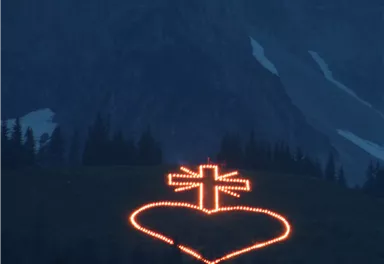 A illuminated heart with a cross in the mountains at dusk. The lights shine in the dark surroundings.