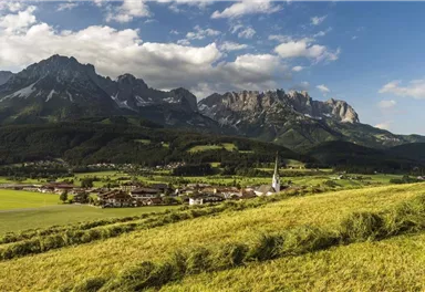 A picturesque landscape with gently rolling hills and a small village. In the background, impressive mountains rise under a blue sky.