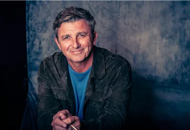 A smiling man with gray-streaked hair sits in front of a neutral background. He wears a jacket and a T-shirt and looks friendly at the camera.