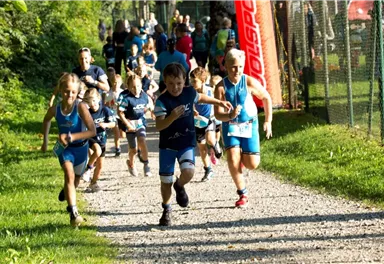 A group of children in sportswear is running on a gravel path. In the background, other participants and a flag can be seen.