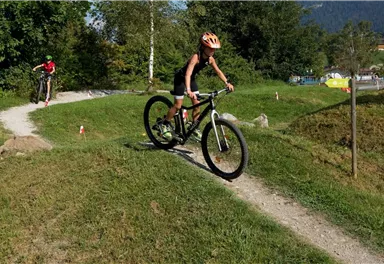 A boy rides a mountain bike over a hilly path. In the background, another cyclist can be seen, surrounded by green landscape.