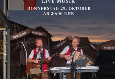 Zwei Musiker treten live auf einer Bühne im Freien auf. Im Hintergrund ist ein schöner Sonnenuntergang und die Berge zu sehen.