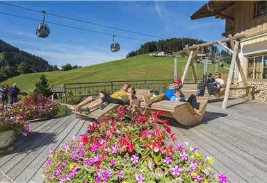 Ein schöner Außenbereich mit bunten Blumen und einer Holzschaukel. Im Hintergrund sind grüne Hügel und Gondeln zu sehen.