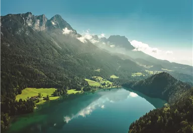 A breathtaking landscape with a clear lake, surrounded by green forests and majestic mountains. The sky is blue and there are some clouds in the distance.