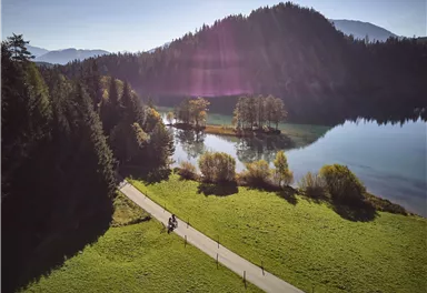Eine malerische Landschaft mit einem ruhigen See, umgeben von Bergen und Bäumen. Ein gepflasterter Weg führt entlang des Wassers, auf dem Radfahrer unterwegs sind.