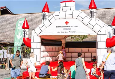 Eine aufblasbare Burg mit roten Türmen steht auf einem Festgelände. Viele Menschen und Kinder haben Spaß beim Spielen und Hüpfen.