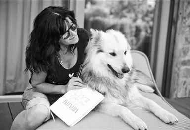 A woman sits relaxed on a lounge chair and strokes her dog. She holds a notebook in her hand and smiles contentedly.