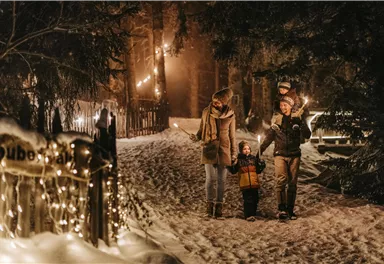 A family walks through a snow-covered forest at night. The path is adorned with lights, creating a festive atmosphere.
