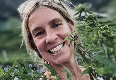 A smiling woman stands among green plants and enjoys nature. In the background, hills and mountains can be seen.
