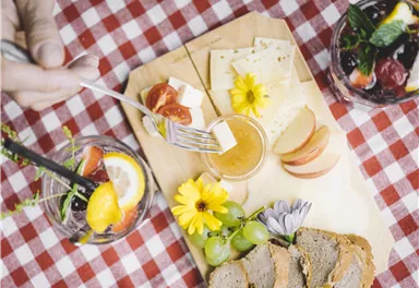 Eine Holzplatte mit Käse, frischem Obst und bunten Blumen. Im Hintergrund stehen zwei Gläser mit Früchte-Cocktails auf einem karierten Tischdeckchen.