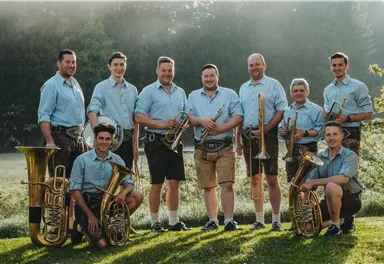 Eine Gruppe von Musikern steht im Freien und trägt traditionelle Trachten. Sie halten verschiedene Blasinstrumente und lächeln in die Kamera.