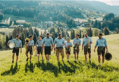 Eine Gruppe von Männern in bayerischer Tracht marschiert über eine grüne Wiese. Im Hintergrund sind Berge und Wälder zu sehen.