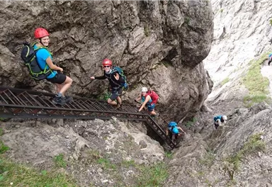 Eine Gruppe von Menschen klettert eine felsige Steigung, während sie einen Helm trägt. Im Hintergrund sind weitere Personen sichtbar, die den Weg erklimmen.