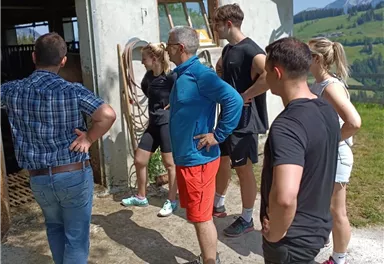 Eine Gruppe von Menschen steht draußen vor einem Bauernhof. Sie schauen sich neugierig etwas an und scheinen interessiert zu sein.