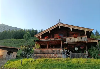 Ein traditionelles Holzhaus in den Bergen, umgeben von grünen Wiesen. Vor dem Balkon sind bunte Blumen und eine Kreuzdekoration zu sehen.