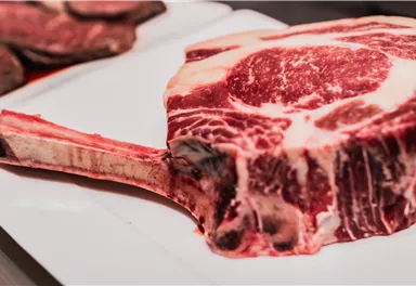 A large, raw tomahawk steak on a white plate. The meat has pronounced marbling and a long bone handle.