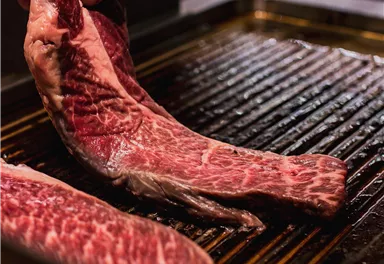 A cook is lifting a piece of red meat onto a grill. The pieces of meat show a nice marbling.