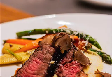 A plate with tender beef, served on creamy mashed potatoes. Next to it are fresh vegetables and a dark sauce.