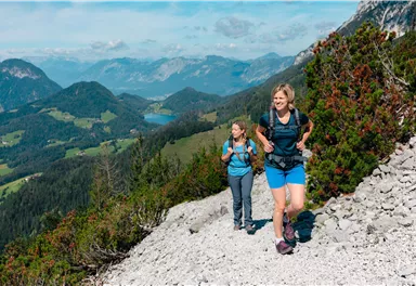 Zwei Frauen wandern auf einem schmalen Pfad in den Bergen. Im Hintergrund erstreckt sich eine malerische Landschaft mit Bergen und Tälern.