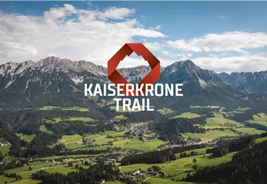 Eine beeindruckende Berglandschaft mit grünen Wiesen und hohen Bergen im Hintergrund. Im Vordergrund steht das Logo für den Kaiserkronen-Trail.