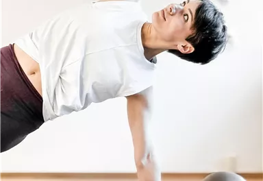 An athletic woman is performing a side plank exercise on a yoga mat. Next to her lies a fitness ball.
