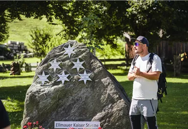 A large stone with five silver stars and a plaque that shows "Ellmauer Kaiser Sterne". Surrounded by colorful flowers and green grass.