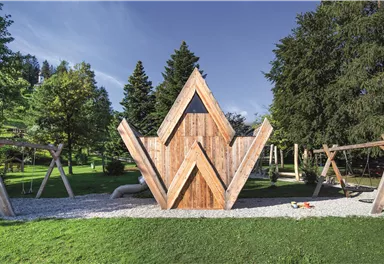 Ein moderner Spielplatz aus Holz mit einem auffälligen, dreieckigen Spielhaus. Im Hintergrund sind Bäume und Schaukeln zu sehen.