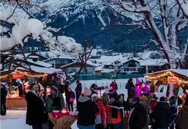 Ein winterlicher Weihnachtsmarkt mit Menschen in warmen Kleidern. Im Hintergrund sind schneebedeckte Berge zu sehen.